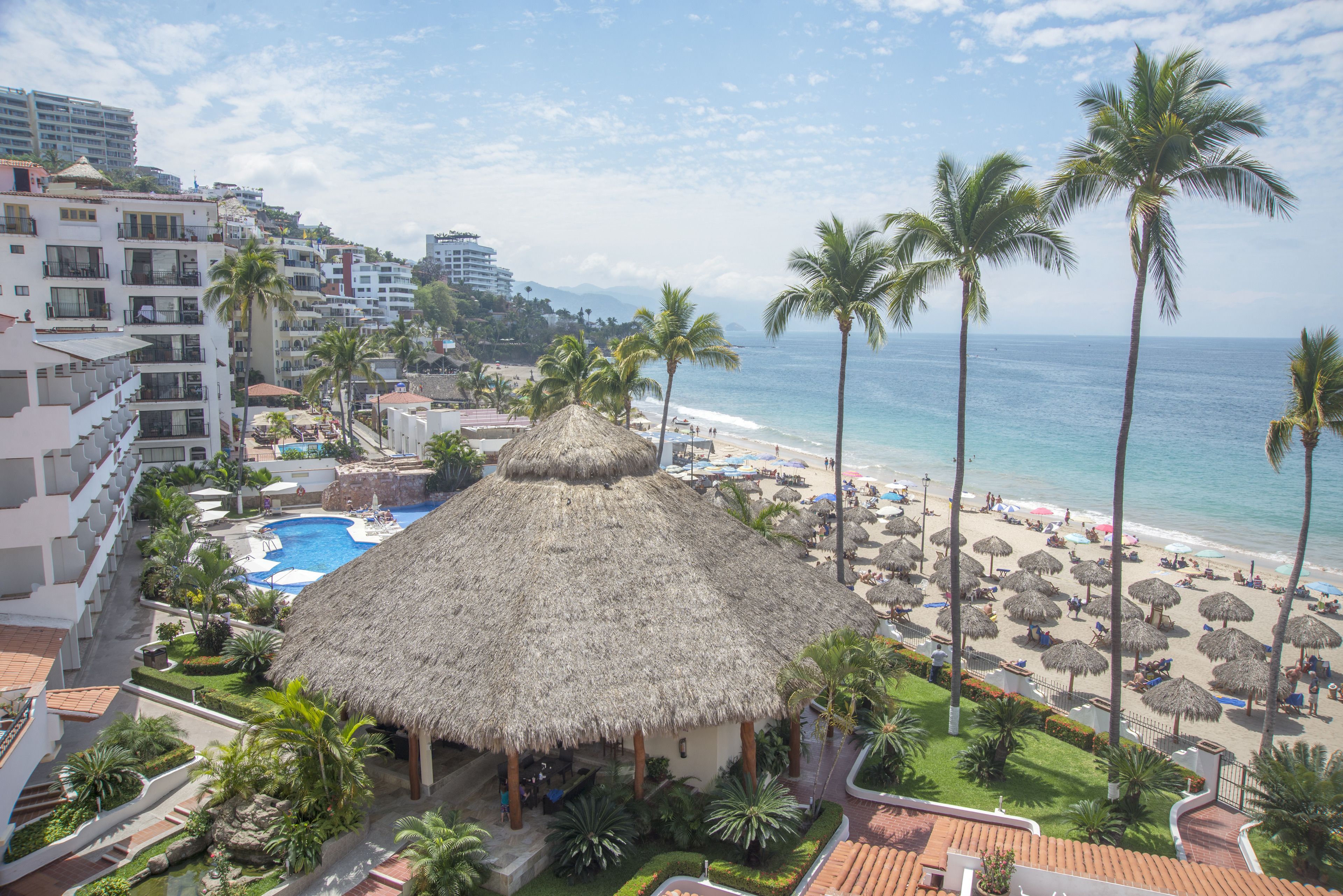 A resort with a pool, thatched-roof structures, palm trees, and a beach with umbrellas.