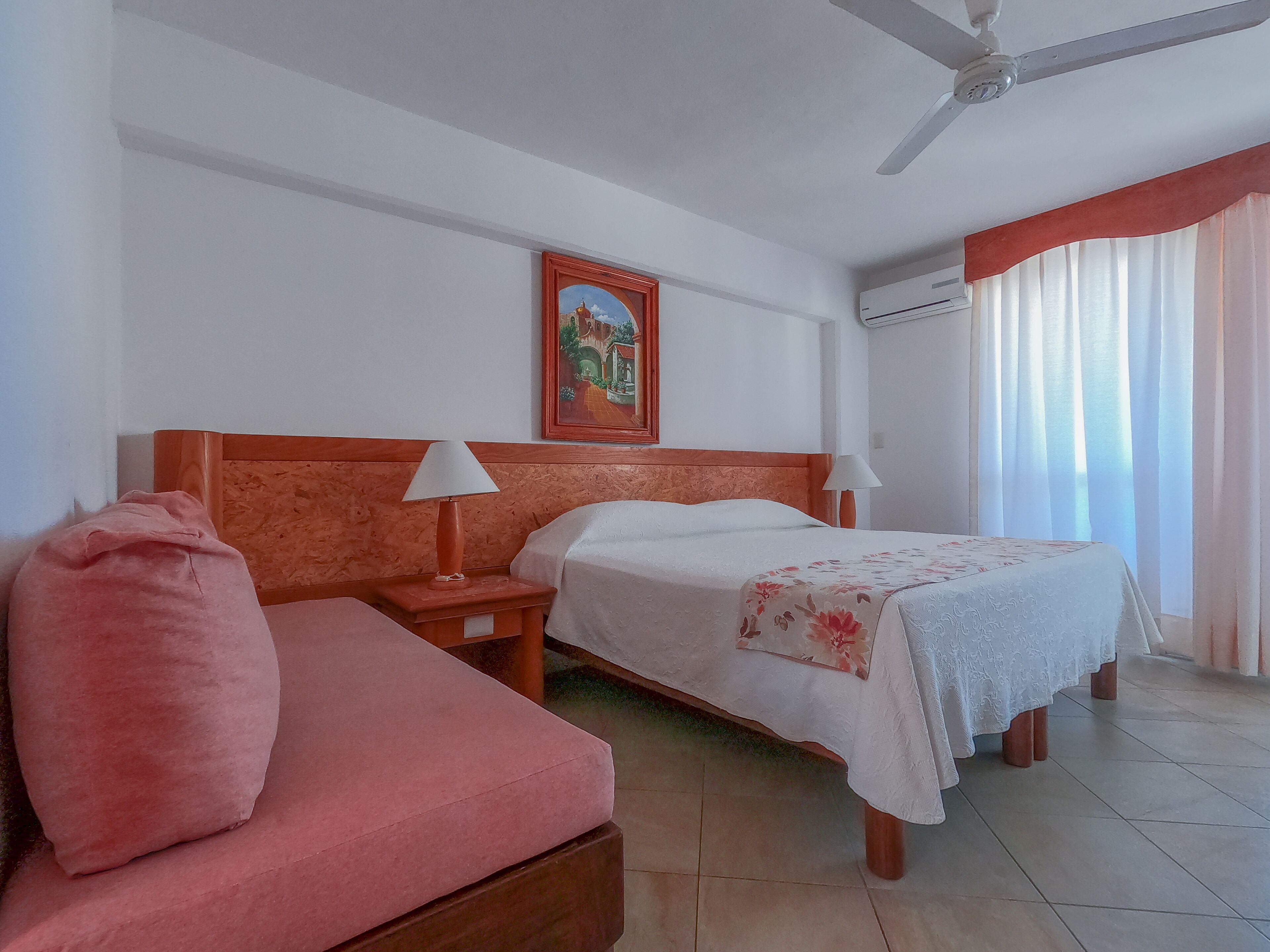 Ein Hotelzimmer mit einem Bett, einem Nachttisch, einer Lampe, einem Deckenventilator, einem Bild an der Wand und einem Fenster mit Vorhängen.