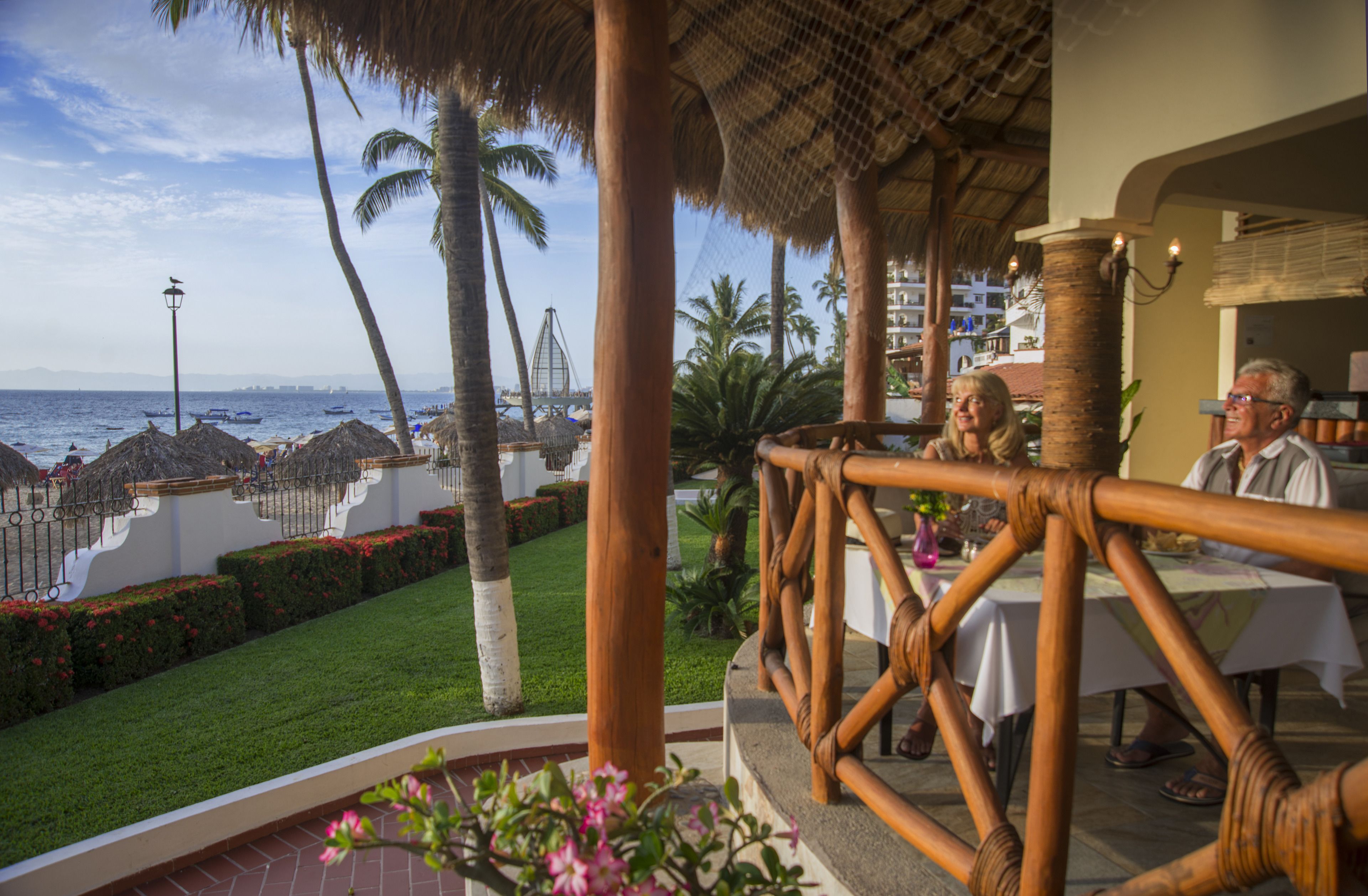 A restaurant with a view of the beach and ocean, featuring a thatched roof and bamboo railing.