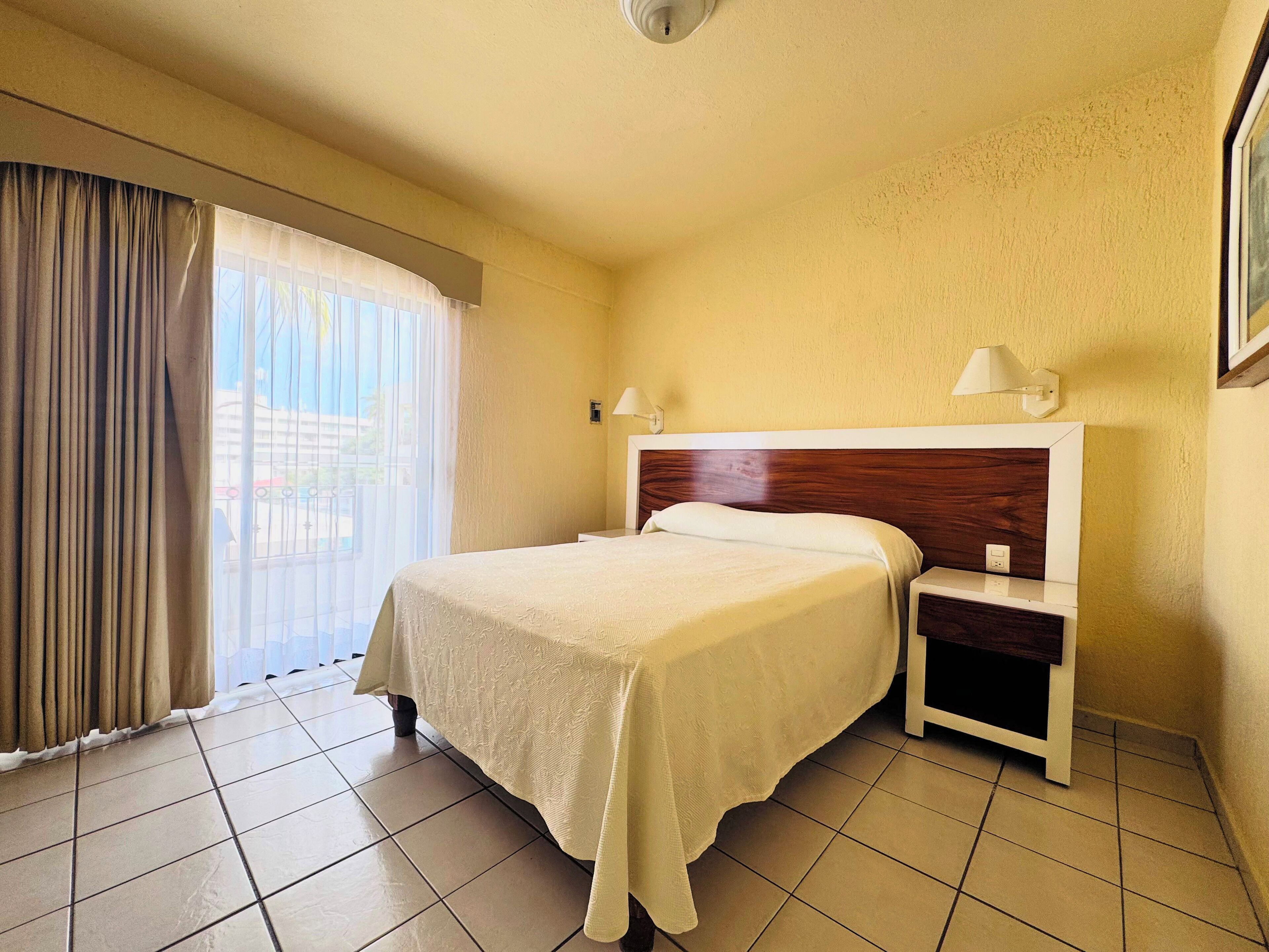 Ein Hotelzimmer mit Bett, Nachttisch, Lampe und Blick durch das Fenster.