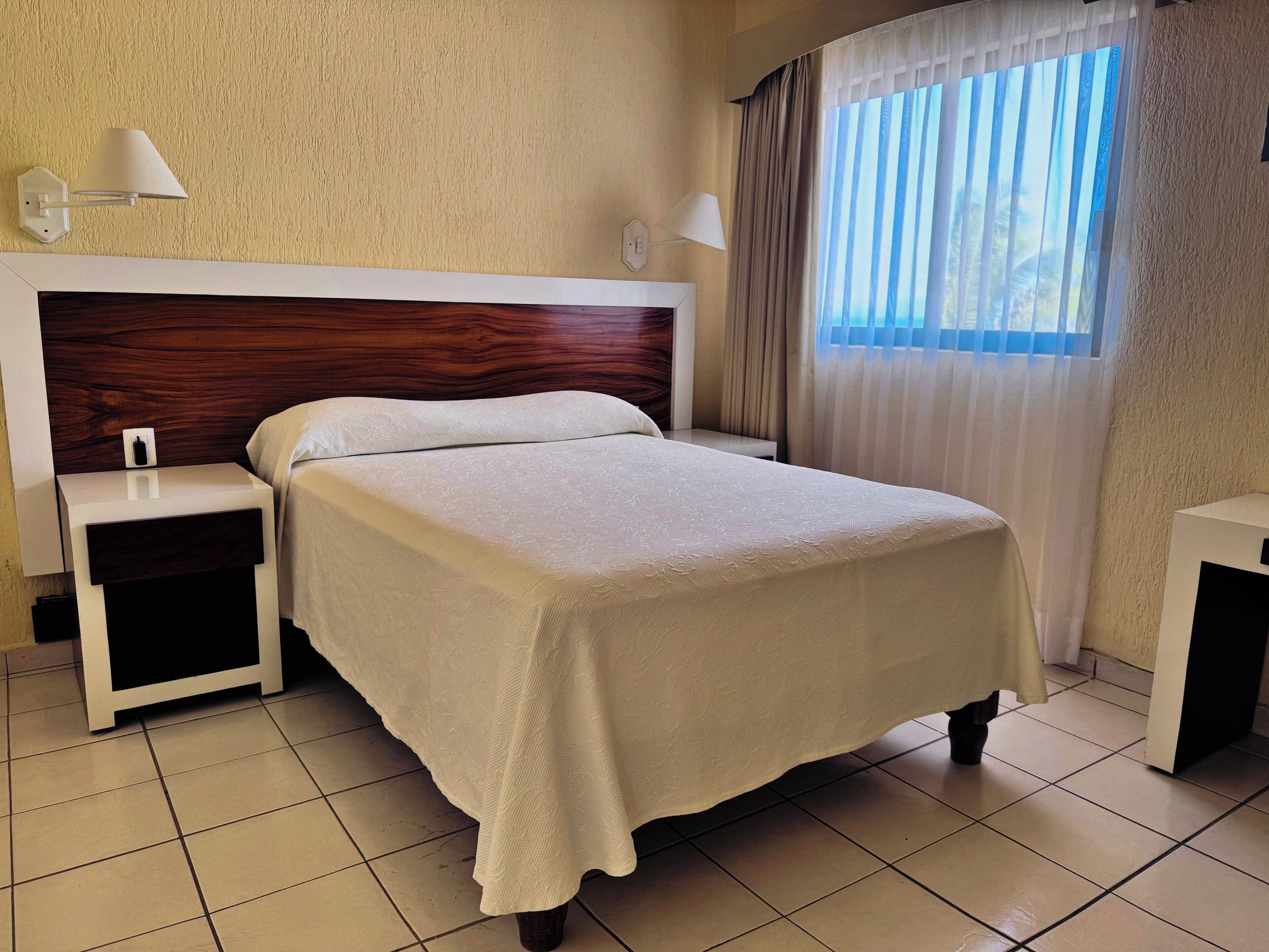 A neatly made bed with a wooden headboard, a bedside table with a lamp, and a window with curtains.