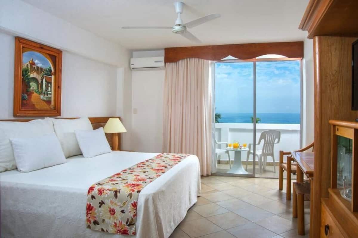 Habitación de hotel con cama, televisor, balcón con mesa y sillas, y un cuadro en la pared.