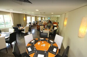 A hotel dining area with round tables, chairs, and a television screen. - Plaza Rio Residences - Barra First (Rio de Janeiro)