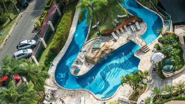 An aerial view of a resort with a large, winding swimming pool surrounded by palm trees and lounge chairs. - Plaza Rio Residences - Barra First (Rio de Janeiro)