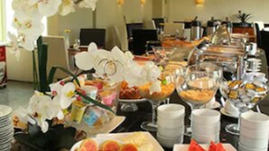 A buffet with various food items, including fruits and desserts, arranged on a table in a well-lit room with a ceiling fan and a large window. - Plaza Rio Residences - Barra First (Rio de Janeiro)