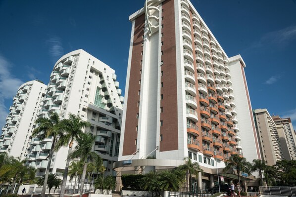 A high-rise hotel with multiple balconies, a prominent central tower, and palm trees in the foreground. - Plaza Rio Residences - Barra First (Rio de Janeiro)