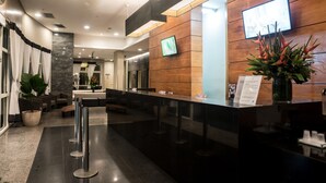 A modern reception area with a wooden reception desk, a flat-screen TV, and a potted plant. - Plaza Rio Residences - Barra First (Rio de Janeiro)