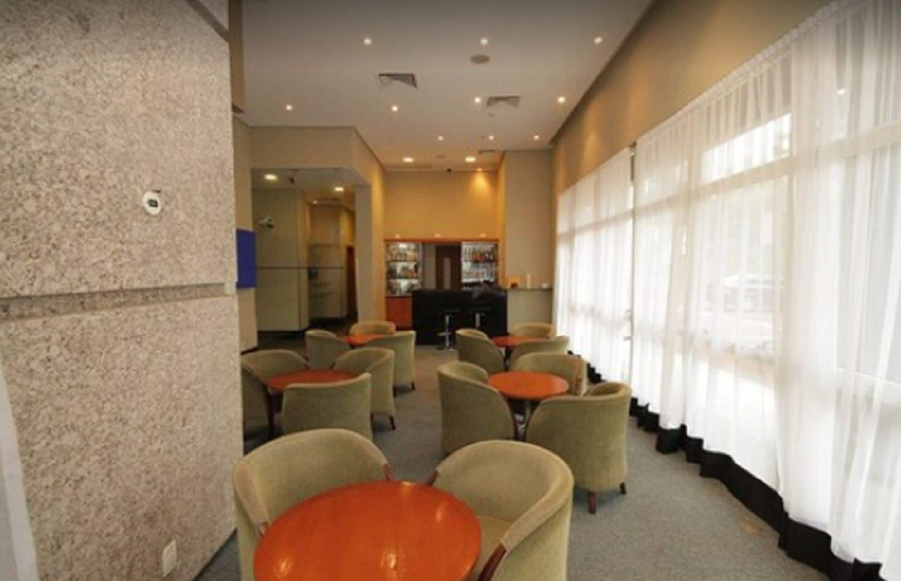 A hotel lobby with multiple round tables and chairs, a bar area, and large windows with sheer curtains.