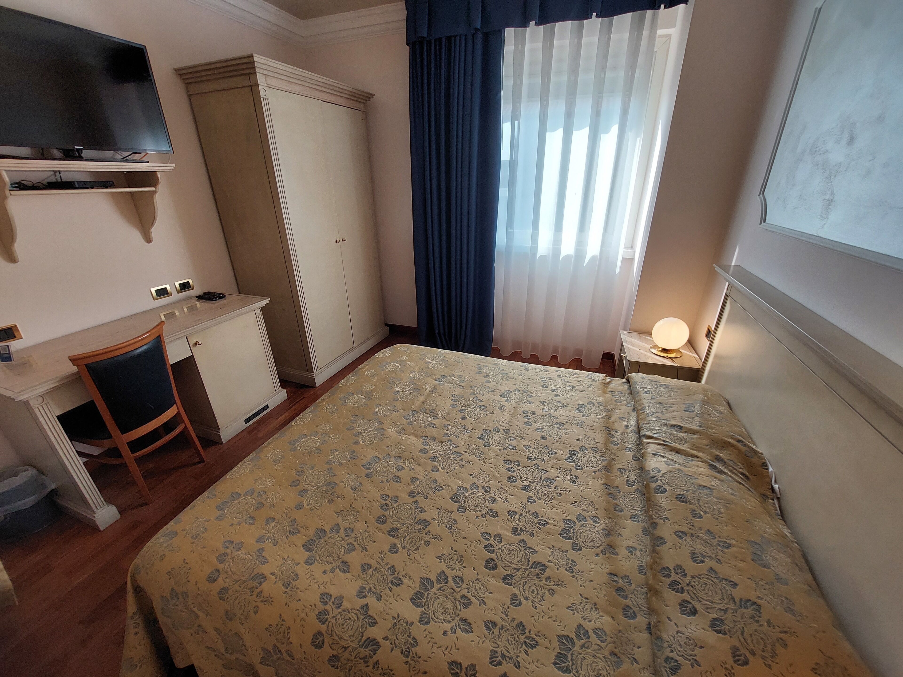 Habitación de hotel con cama, escritorio, silla, armario, televisor y ventana con cortinas.
