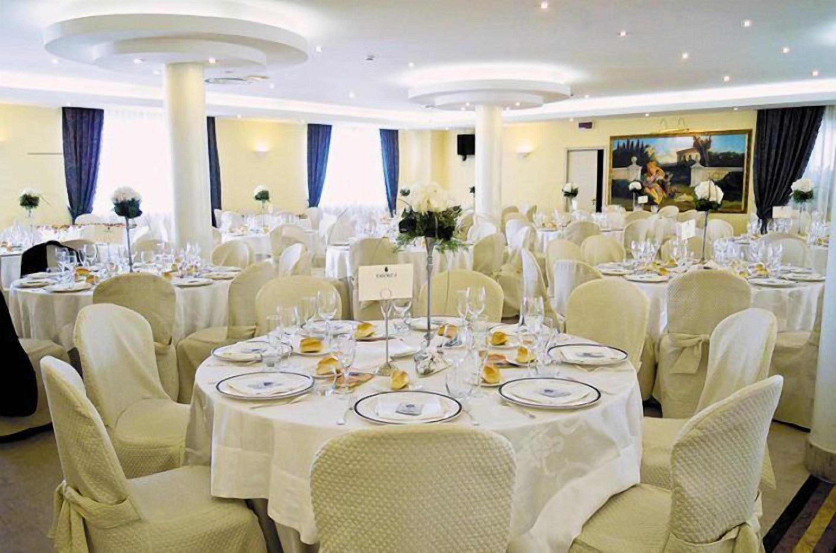 Un salón de banquetes con mesas redondas dispuestas para comer, sillas blancas y un cuadro grande en la pared.