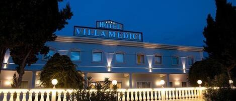 A hotel named "Hotel Villa Medici" with a classical facade, illuminated windows, and a well-maintained garden.