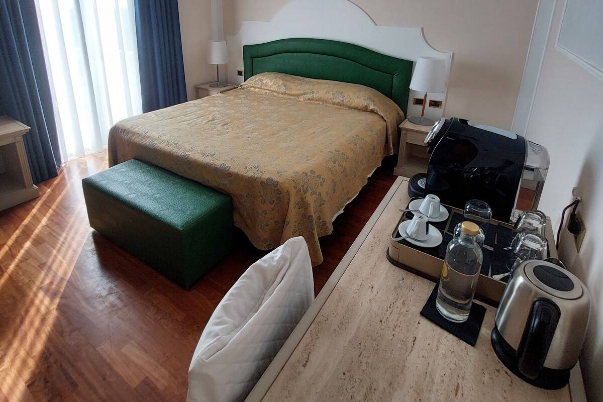 A hotel room with a bed, a desk with a coffee maker, a kettle, and a remote control.