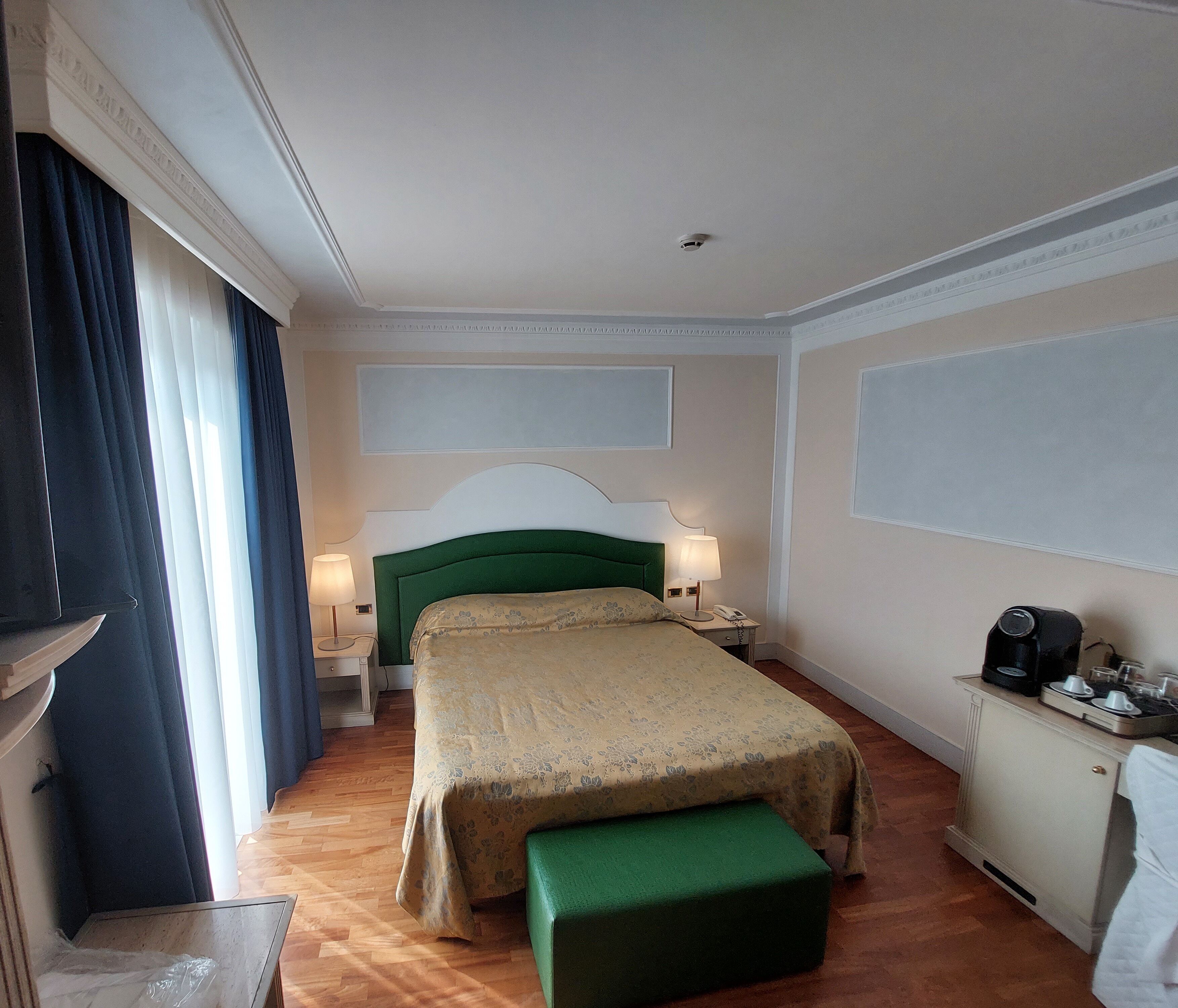 Una camera d'albergo con un letto, comodini, una scrivania con una macchina per il caffè e una finestra con tende.