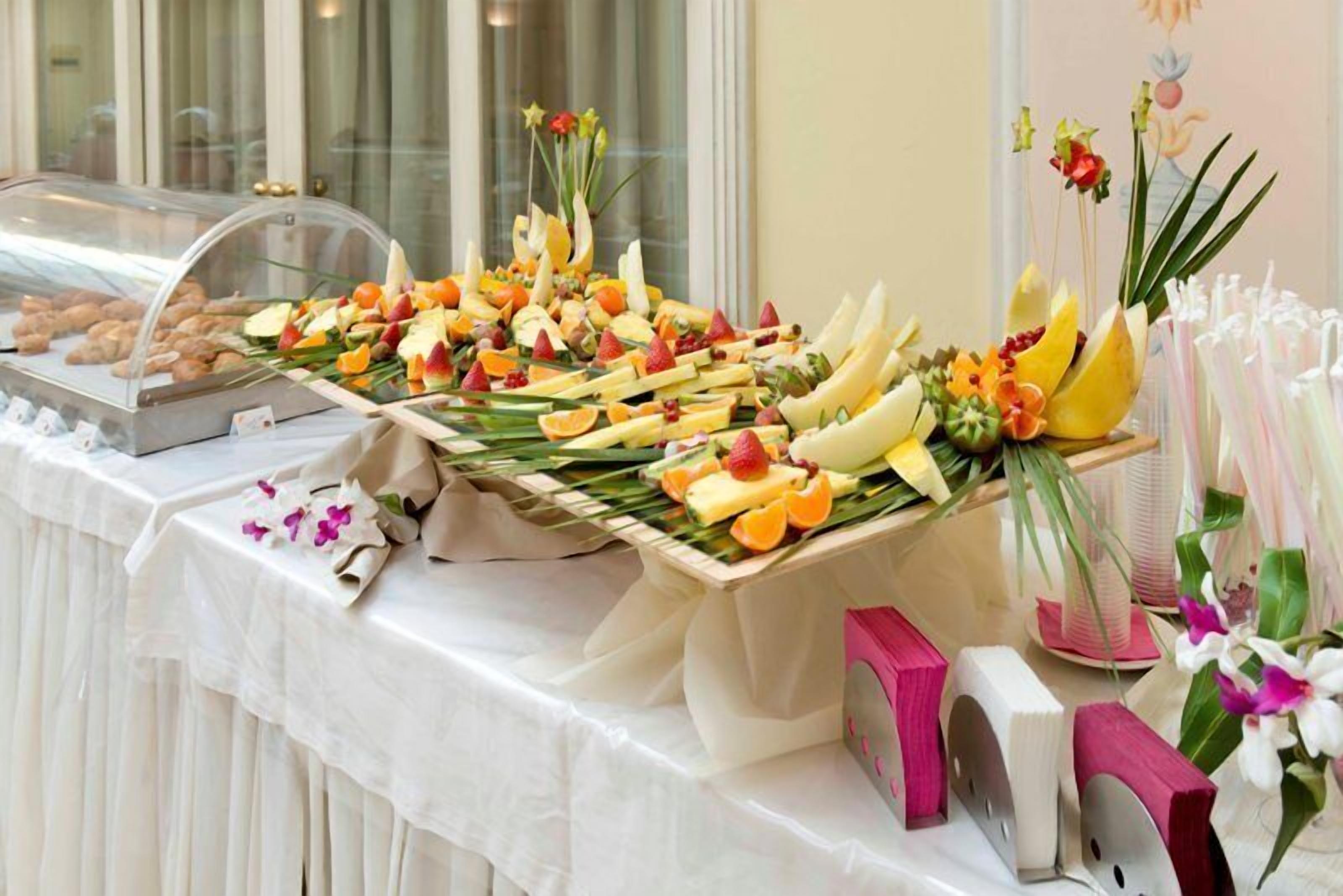 Een buffet met diverse soorten fruit en gebak, een glazen vitrine met gebak, en een decoratieve bloemenstelling.