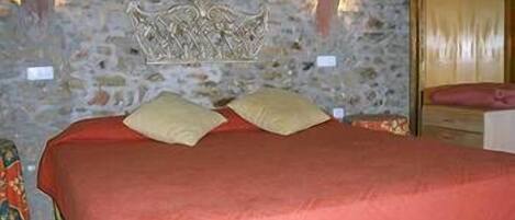 Una cama con una colcha roja y dos almohadas, una pared de piedra con un escudo decorativo, y una puerta de madera con marco dorado.