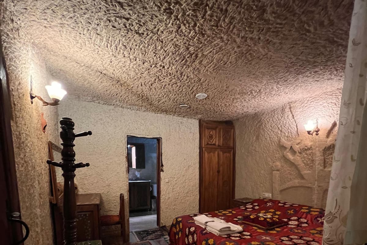 Ein Zimmer mit strukturierter Decke, einem Bett mit gemustertem Überwurf, einem Holzschrank und einem kleinen Tisch mit Lampe.