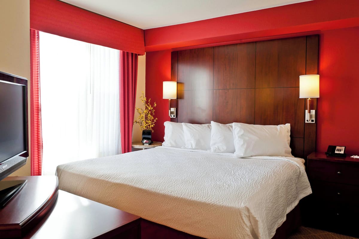 Ein Hotelzimmer mit einem großen Bett, zwei Nachttischlampen, einem Fernseher und einer Vase mit Blumen.