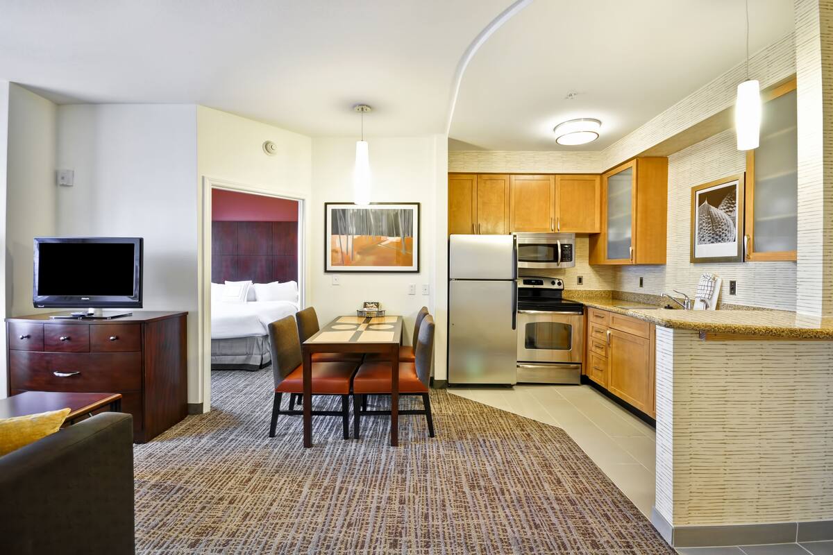 Ein Hotelzimmer mit einem Bett, einem Fernseher, einem Essbereich mit Tisch und Stühlen sowie einer Küchenzeile mit Kühlschrank, Backofen und Mikrowelle.