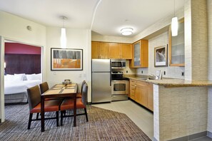 A compact hotel room with a kitchenette, a dining area with a table and chairs, and a bed with a red headboard.