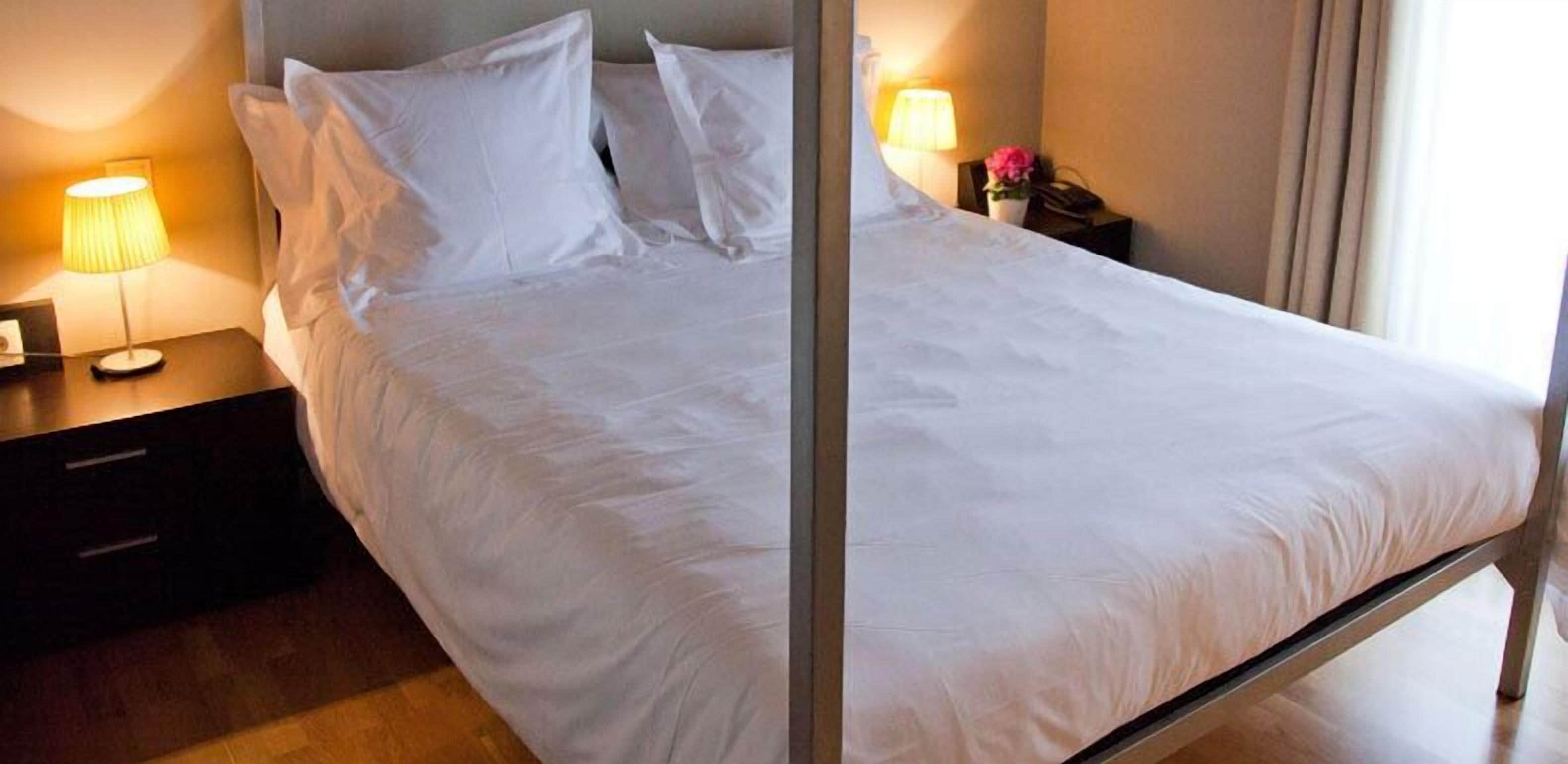 A neatly made bed with white linens, a bedside table with a lamp, and a nightstand with a phone and a small vase of flowers.