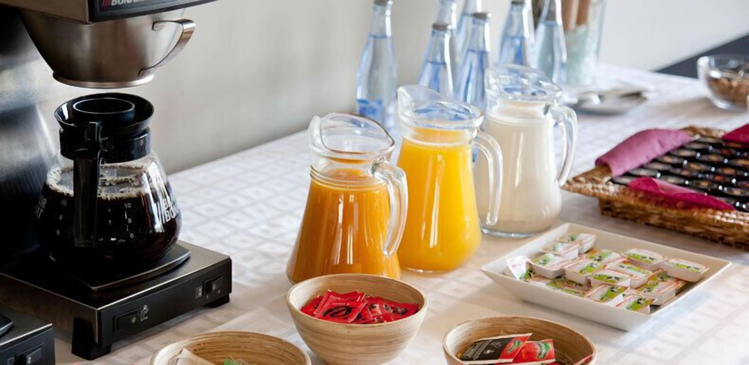 Eine Kaffeemaschine, zwei Orangensaftkaraffen, eine Milchkrug und eine Auswahl an Backwaren auf einem Tisch.