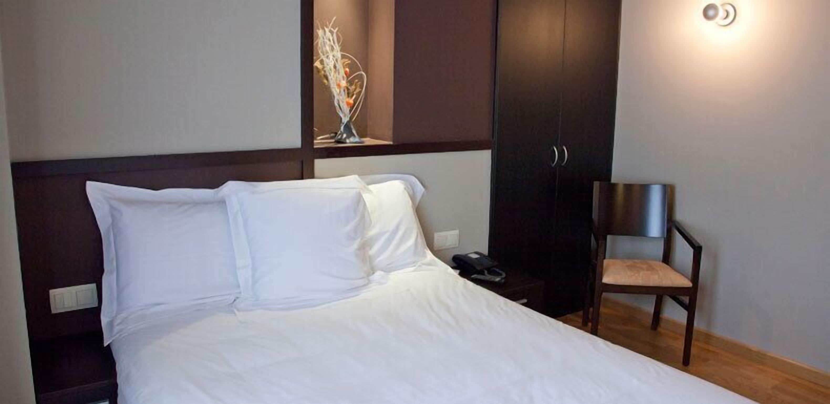 A neatly made bed with white linens, a wooden nightstand, a chair, and a dark wardrobe.