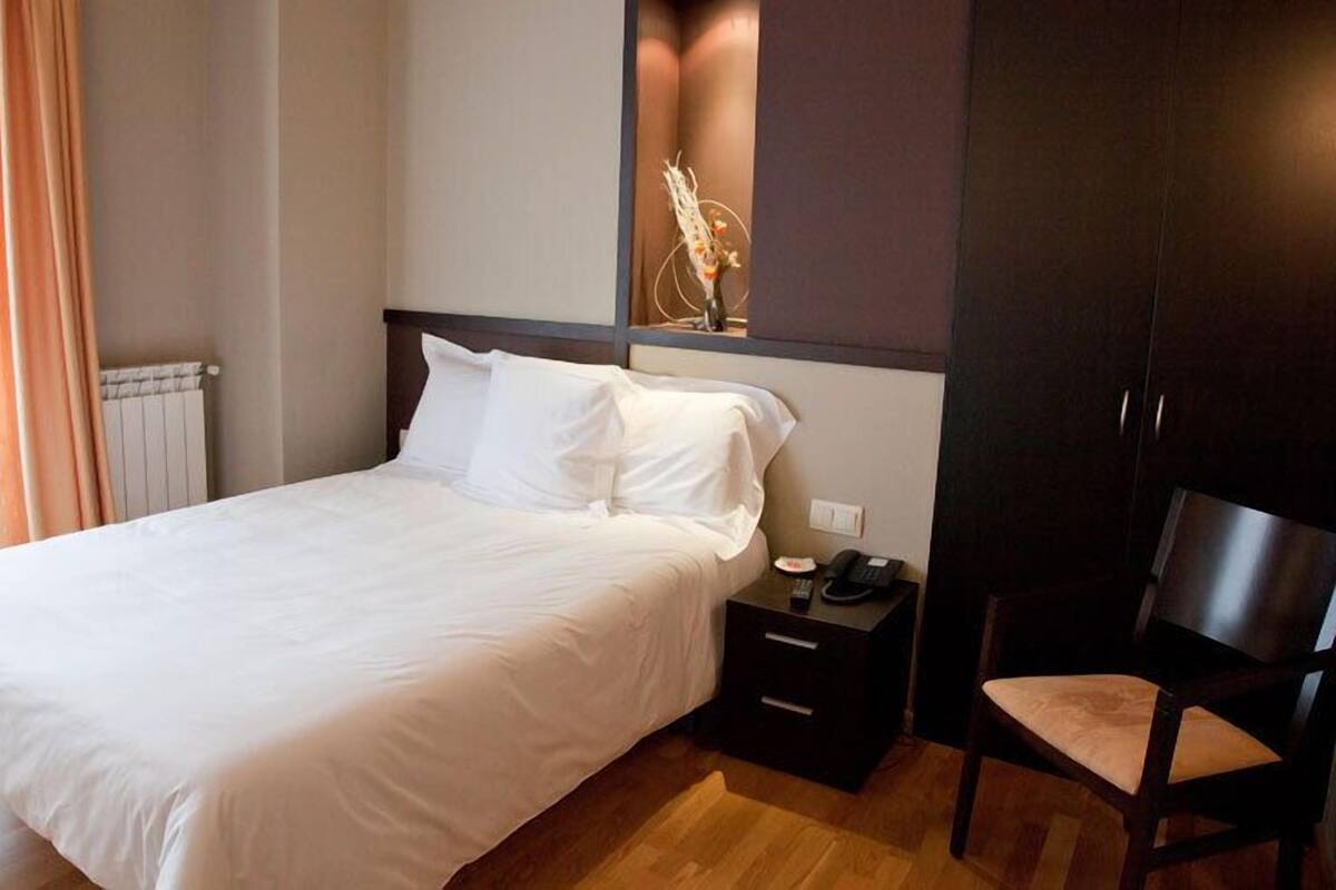 Un lit bien fait, avec du linge de lit blanc, une table de chevet avec un téléphone et une lampe, et une chaise avec un coussin.