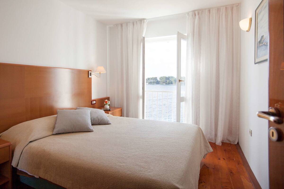 A hotel room with a large bed, a wooden headboard, a nightstand with a lamp, and a view of the water through the window.