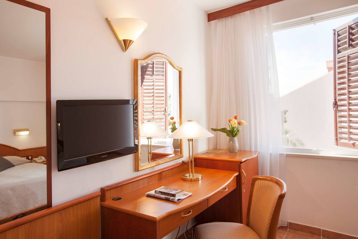 A hotel room with a bed, a desk with a lamp, a television, a mirror, and a window with blinds.