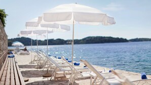 A seaside promenade with white lounge chairs and umbrellas, a stone pathway, and a view of the sea and distant land. - Hotel Odisej (Mljet)