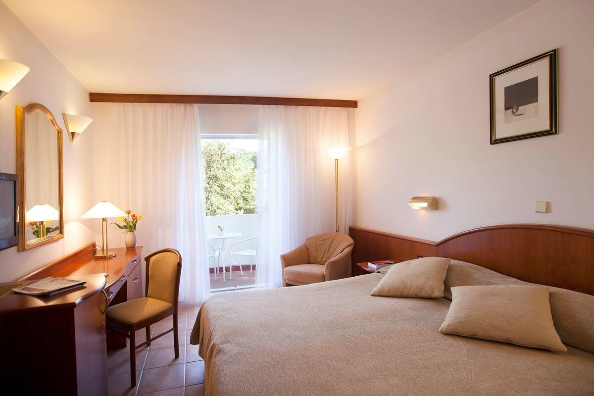 A hotel room with a bed, a desk, a chair, a lamp, and a window with curtains.