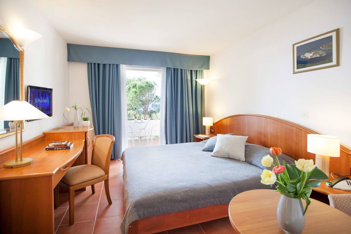 A hotel room with a bed, desk, chair, and a small table with a vase of flowers.