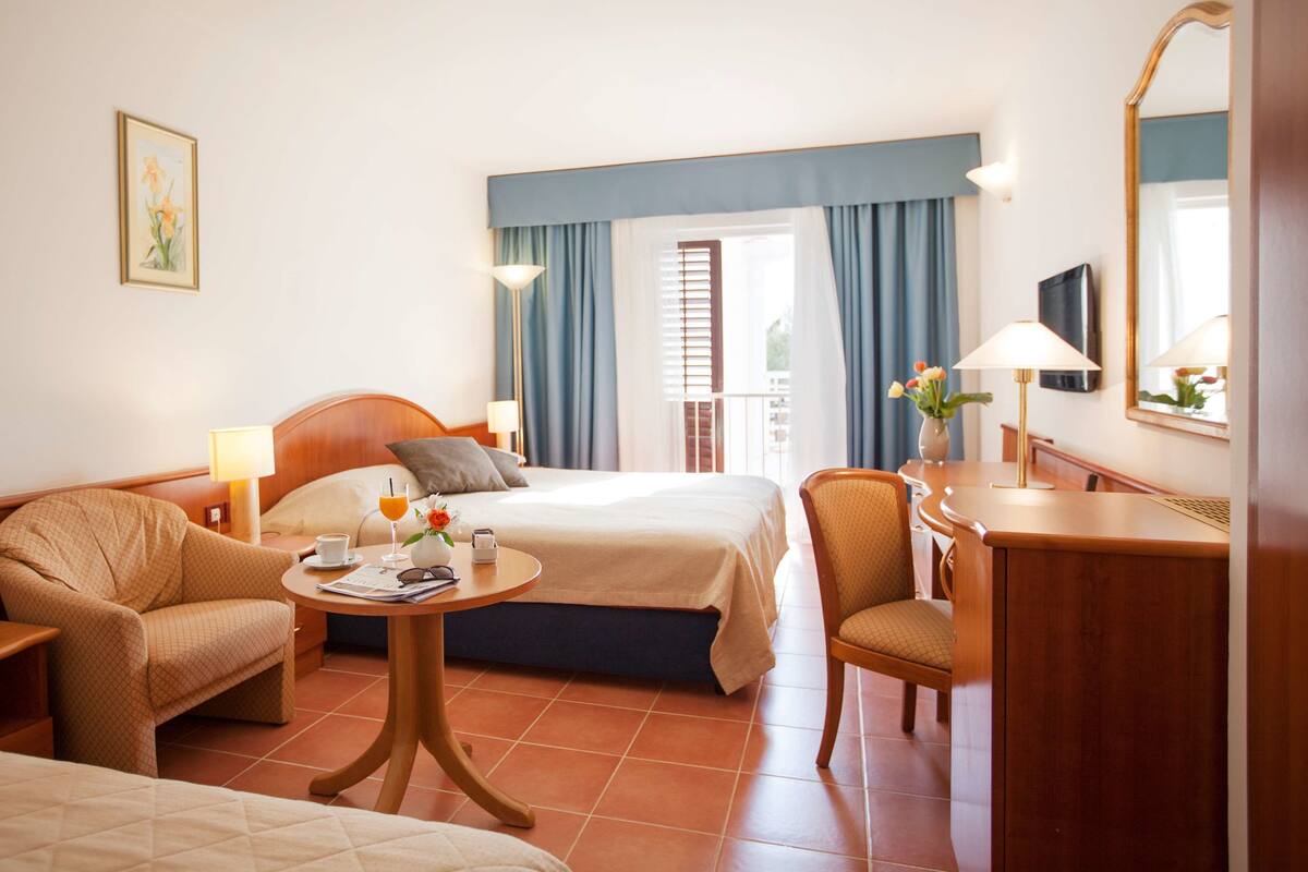 A hotel room with a bed, two chairs, a desk, a lamp, a TV, and a window with curtains.