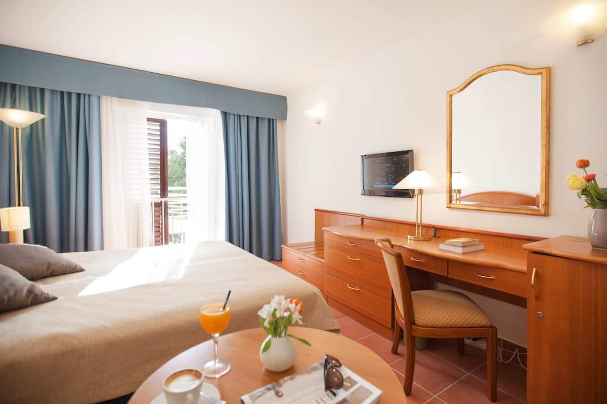 A hotel room with a bed, a desk, a chair, a table with a drink and flowers, and a TV on the wall.