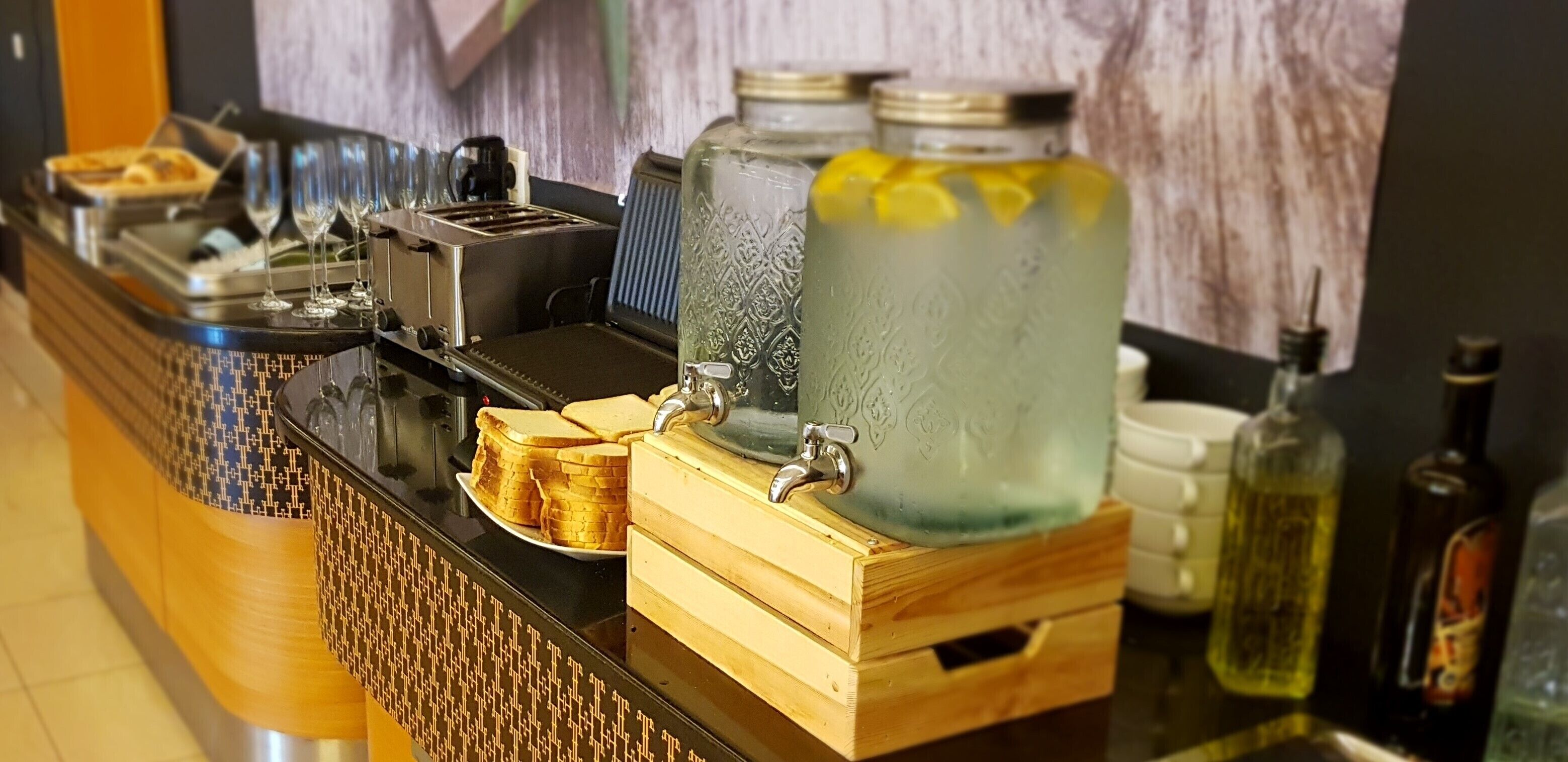 A bar area with a coffee machine, glasses, a tray of bread, and large water dispensers.