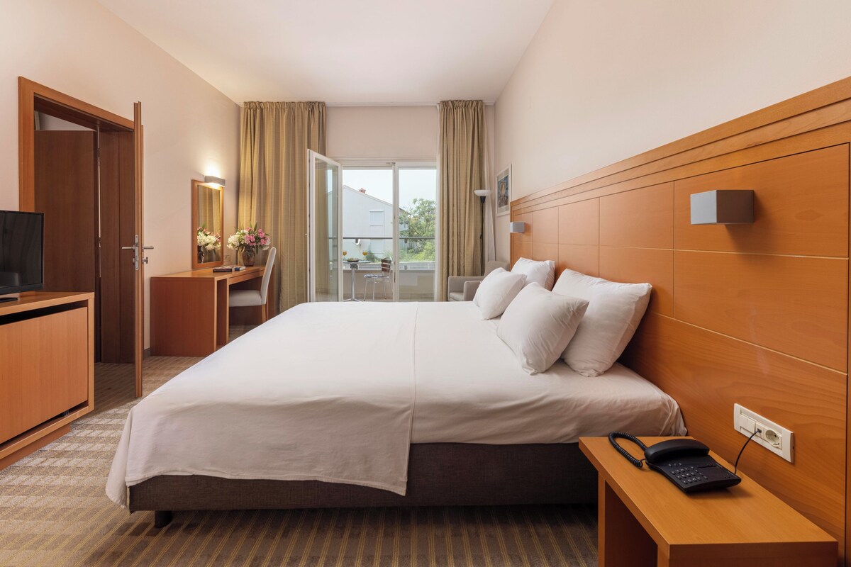 A hotel room with a bed, a desk with a TV, a bedside table with a phone, and a view of the outside.