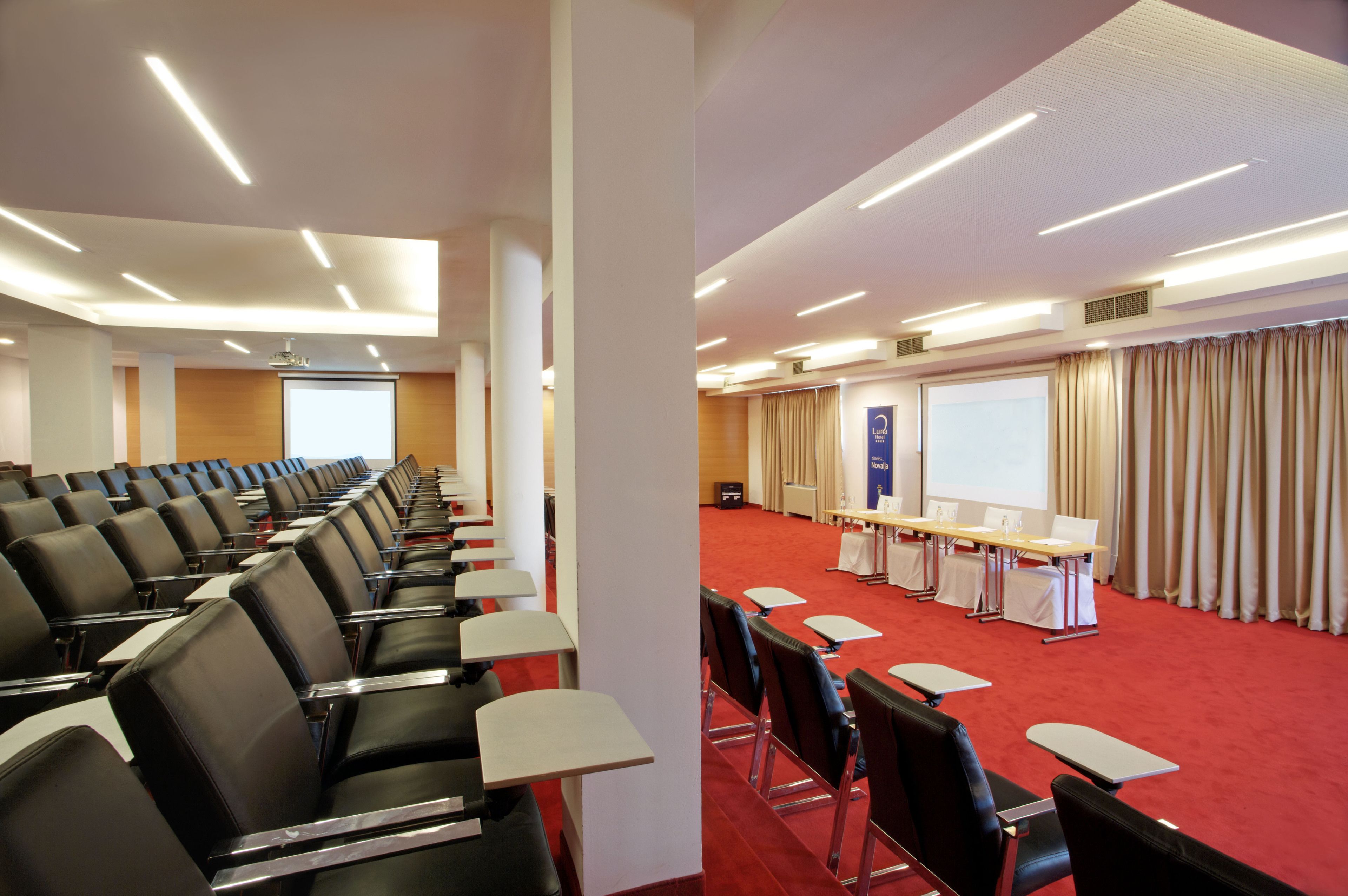 Un salón de conferencias con filas de sillas negras, una alfombra roja, una pantalla de proyector y un atril.