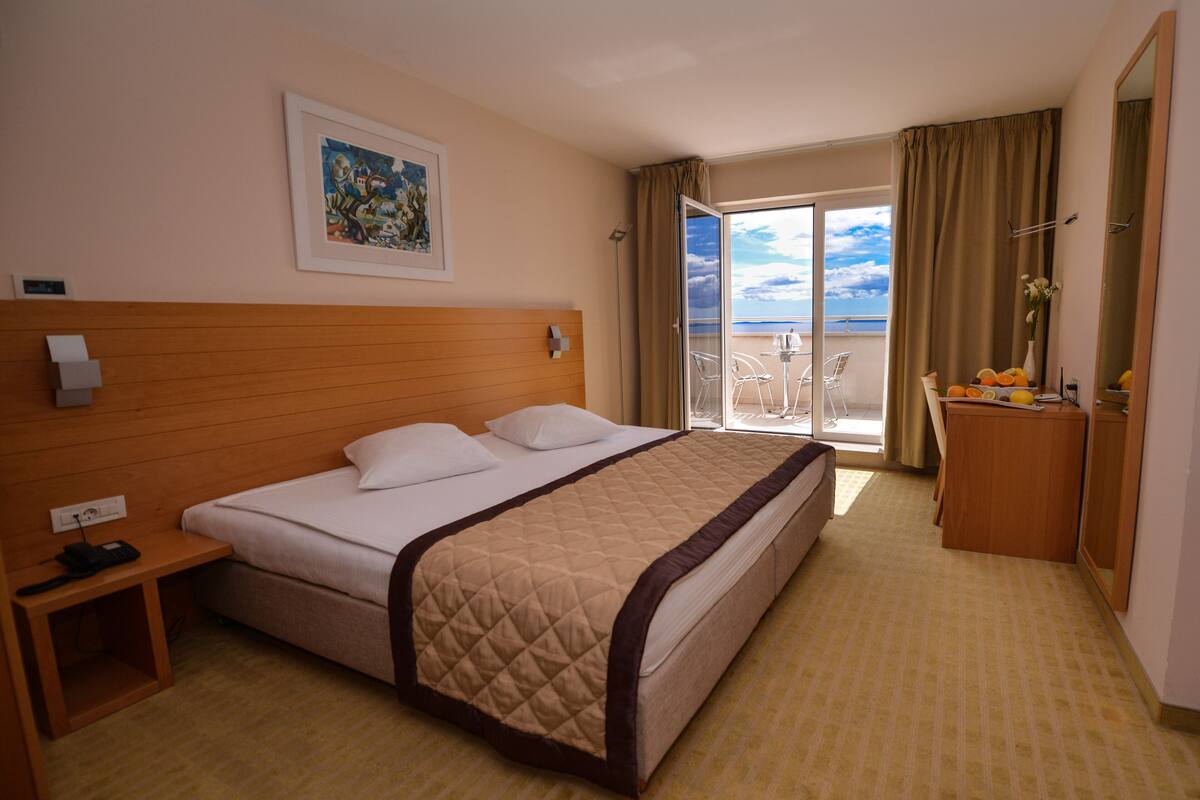 A hotel room with a large bed, a bedside table with a phone, a painting on the wall, and a view of the balcony.