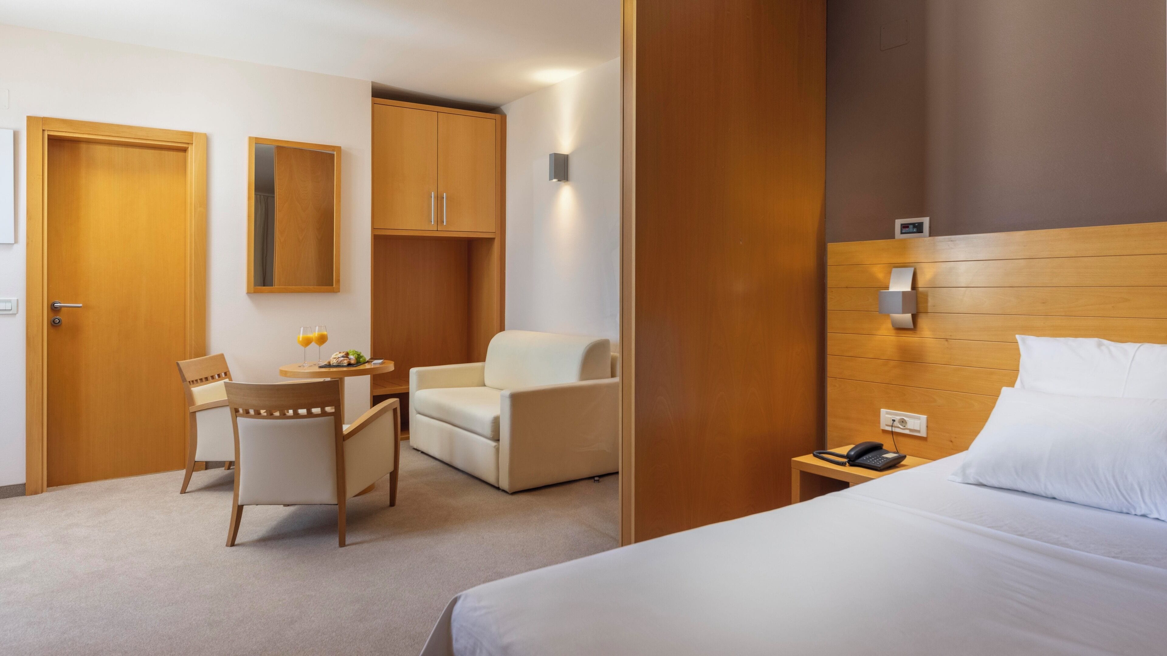 Habitación de hotel con cama, escritorio y dos sillas, una mesita con dos vasos de zumo de naranja, y un espejo en la pared.