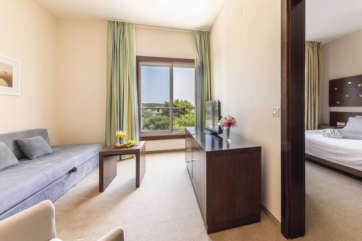 A hotel room with a bed, a sofa, a desk with a TV, and a window with curtains.