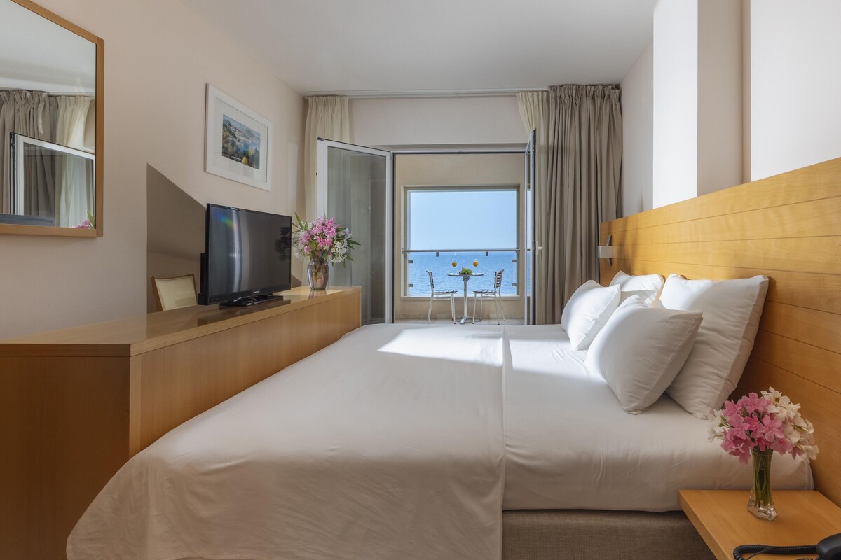 A hotel room with a large bed, a TV on a wooden desk, a small table with drinks, a vase of flowers, and a view of the sea through a balcony.
