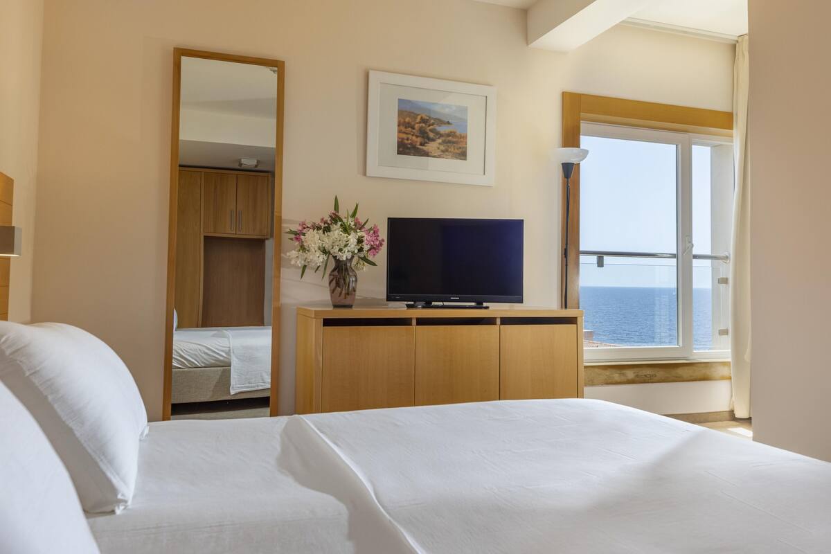 A hotel room with two beds, a TV on a wooden cabinet, a mirror, a painting, and a view of the sea through a large window.