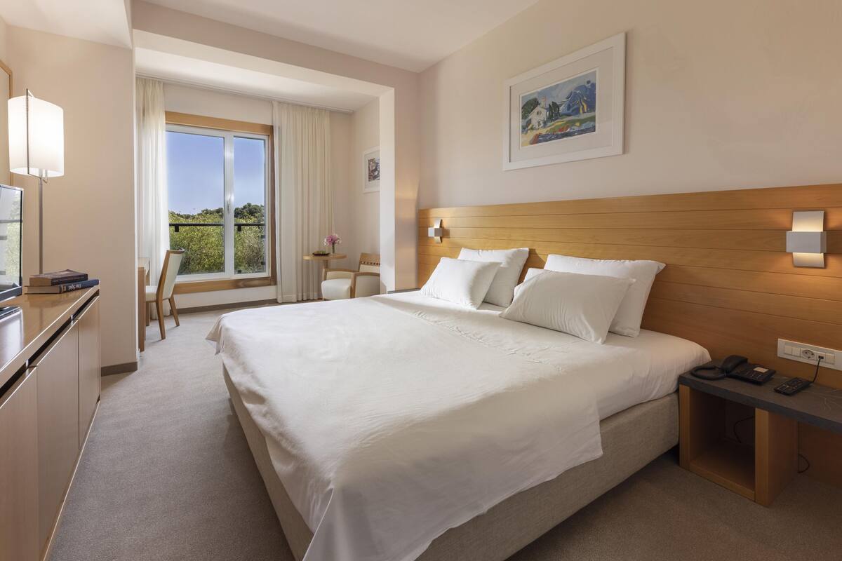 A hotel room with a large bed, a desk with a TV, a chair, a small table with a vase, and a painting on the wall.