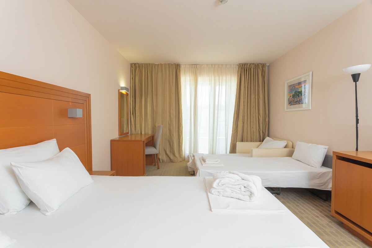 A hotel room with two beds, a desk, a chair, and a lamp.