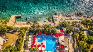 An aerial view of a resort with a swimming pool surrounded by red umbrellas, a beach area with lounge chairs, and a rocky shoreline. - Family Hotel La Luna (Novalja)