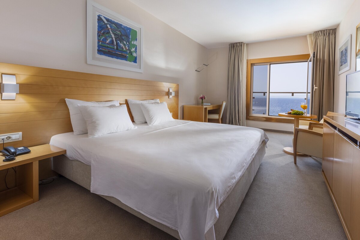 A hotel room with a large bed, a desk with a telephone, a television, and a window with a view of the sea.