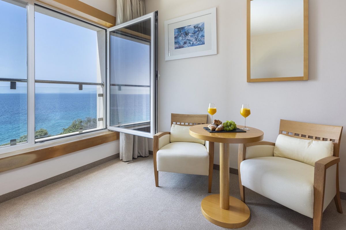 A hotel room with a large window overlooking the sea, two chairs, a small round table with breakfast items, and a framed painting on the wall.