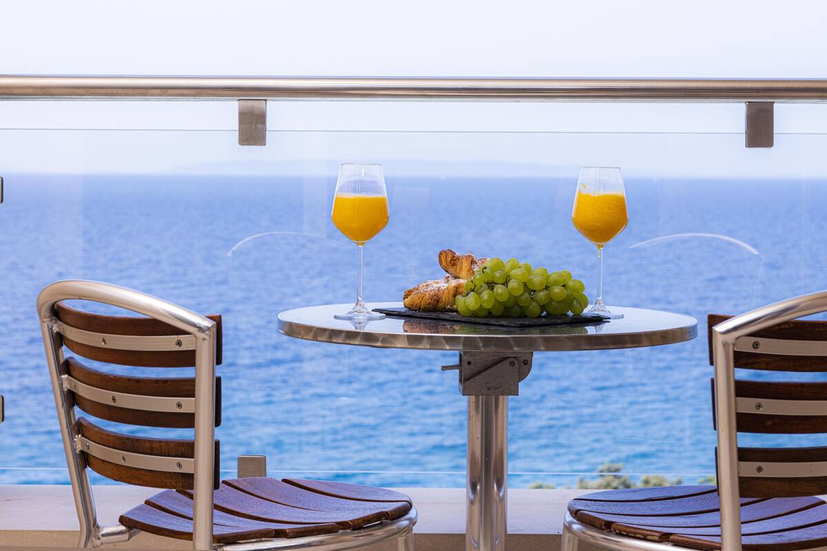 A table with two chairs, a croissant, grapes, and two glasses of orange juice, overlooking the sea.