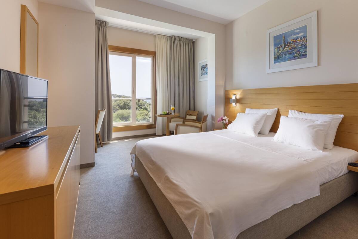 A hotel room with a large bed, a desk with a TV, a chair, a small table with a drink, and a window with curtains.