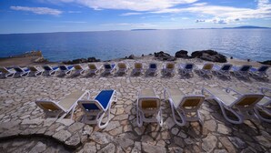 A row of sun loungers on a stone-paved beach with a view of the sea and distant land. - Family Hotel La Luna (Novalja)